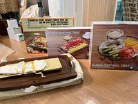 Photo showing boxed stainless steel roast/bake pan set, boxed 3-piece glass serving tray set (two boxes) and wooden meat carving board on floor.