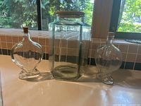 Three decorative glass jars displayed near a window: one large rectangular jar with a wooden lid and two heart-shaped jars with cork stoppers.