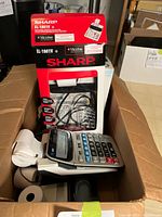 Photo of Sharp EL-1801V electronic printing calculator partially outside original box, with visible paper rolls and power cord inside a cardboard container.