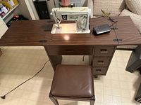 Singer sewing machine set in a brown wooden sewing table with attached chair, power cord visible on floor