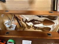 Photo showing a wooden drawer containing carved teak serving utensils with giraffe handles and two metal wine bottle stoppers in boxes, grouped together.
