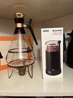 Braun Aromatic coffee grinder in box next to Atomic Inland glass coffee carafe on metal warmer stand