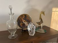 Wide view showing all four main items together on a wood surface: wine decanter, Norman Rockwell plate, glass giraffe, and glass lily paperweight.