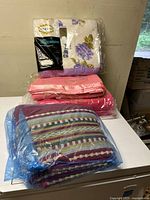 Four folded vintage blankets, one in white floral Magic Weave packaging, stacked on a white surface in a basement area.