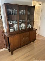 Front view of the vintage teak buffet and hutch showing three glass doors on the hutch top revealing shelving with glassware and china inside and four sliding wooden doors on the buffet below with decorative carved panels on the middle two doors.