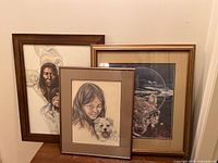 Three framed art prints side by side leaning against wall on floor: Indigenous portrait, girl with dog, nature scene.