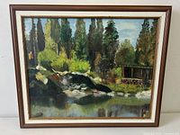 Framed oil painting on canvas of a lake vista scene, showing natural landscape with trees, water, rocks, and a wooden structure. Signed by KUHL.