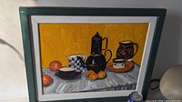 Frontal view of the still life oil painting on board with green frame, showing black coffee pot, checkered bowl, cup with saucer, and oranges on a gray cloth with yellow background.
