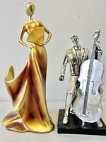 Both the gold-tone female figure and silver-tone male jazz musician sculptures shown together for scale and condition.