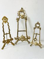 Full view of three vintage brass easels of varying heights arranged side by side against a white wall