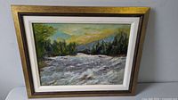 Oil painting of turbulent waters and forested landscape in a gold and white frame, signed by Nicholls.