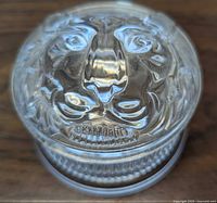 Circular clear art glass ring box with lion head sculpted lid shown from the front.