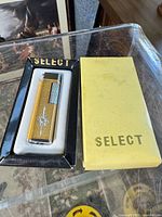 Photo showing the SELECT branded lighter inside the original black plastic insert holder and next to the yellow box with SELECT lettering.