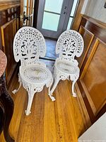 Photo showing two white aluminium garden chairs with intricate decorative patterns on backs and seats placed on wooden flooring indoors near a door.