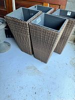Front view of four tall square brown wicker planter plinths with black plastic liners