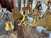 Four vintage brass items displayed on a table: two ship's wheel bells, a seated figure holding a Belfast sign, and an abstract brass shoe form.