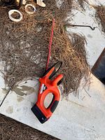 Full view of the Black & Decker cordless hedge cutter lying on concrete with pine needles around.