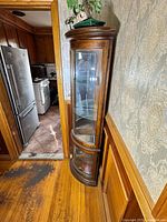 Full view of wooden corner curio cabinet placed in corner showing rounded glass door and shelves.
