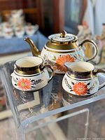 Tea pot, creamer, and sugar dish displayed on clear acrylic stand showing floral and gold design