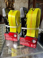 Two new heavy duty Husky ratcheting tie downs with yellow straps and metal hooks in original red boxes with white bands securing the straps.