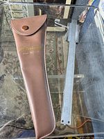 Full view of the mitutoyo vernier caliper beside the brown protective case with button flap, both lying on a glass surface.