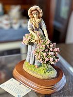Front view of the hand painted country folk lady figurine with her bonnet, dress, and pink flowers, showing wooden base and grass detail.