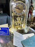 Front view of the Brass Birks anniversary clock showing dial, pendulum balls, and domed glass cover with some small included paperwork or manuals near base.