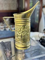 Full view of the embossed brass pitcher showing its tall shape, wide spout and large angular handle.
