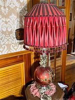 Side view of one lamp showing the red spherical base with gold floral design, silver ornate stand, and original pleated red and black striped shade with hanging tassels.