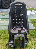 Front view of the black child bike seat showing the adjustable shoulder straps, waist buckle, and footrests with straps on a grassy background.