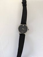 Front view of the black vintage woman's wristwatch with silver round case, black dial with single dot marker at 12 o'clock, and black textured leather strap.