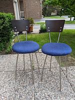 Front view of both blue fabric upholstered bar stools with metal legs and dark wooden backrests seen outdoors on pavement.