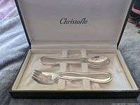 Opened box showing silver baby spoon and fork inside white fabric lining with holding straps.