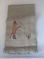 Full view of folded beige linen tea towel with embroidered fox and monkey holding sparklers.