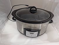 Front angled view of the Crock Pot showing stainless steel body, clear glass lid with black handle, digital control panel and power cord.