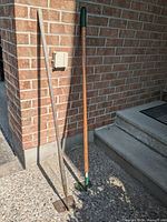 Full view of two garden hoes leaning against a brick wall near steps, showing both wooden and metal handles with attached blades.