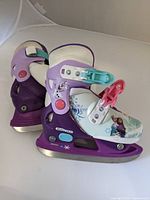 Pair of purple and white Frozen-themed kids ice skates with adjustable sizing shown from an angle highlighting the graphics and blade.