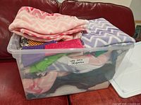 Large plastic bin filled with assorted decorative fabric pieces, mainly fleece on top in pink and lavender chevron patterns.