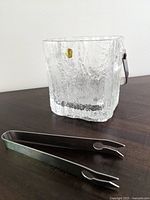 Clear textured glass ice bucket with metal handle and metal ice tongs placed on dark surface