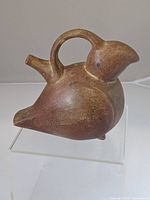Front and side view of the matte brown ceramic bird figure with a handle arch and spout, displayed on a clear stand.