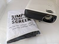 Projector angled showing lens and original packaged simple projection screen labeled 100 inch