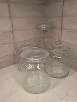 Four clear glass storage containers with lids displayed on kitchen counter against tiled backsplash. Three shorter containers and one taller container.
