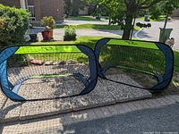 Two portable soccer nets set up on ground outside on stone tiles, showing black mesh, green top and bottom strips, and blue edges.