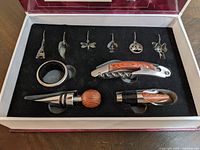 Overhead view of wine kit items arranged in black foam insert inside gift box, showing corkscrew, bottle stoppers, wine pourer, foil cutter, and six decorative wine glass charms.