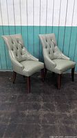 Two grey tufted pleather side chairs with dark wooden legs against white and teal paneled background