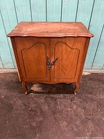 Frontal view of the two door Louis XV style wood cabinet standing against a pale blue wooden panel wall on a concrete floor.