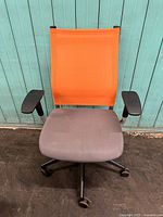 Front view of the orange mesh backrest and grey upholstered seat with black armrests on caster base.