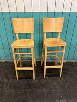 Front view of two maple wood barstools with backs showing worn seats and surface scratches.