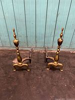 Full view of both brass and iron andirons together against a vertical wood panel background, showing their paired design and size.