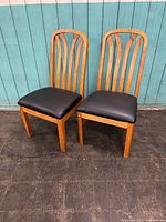 Pair of solid oak dining chairs with black leather seats, front view against teal paneled wall.